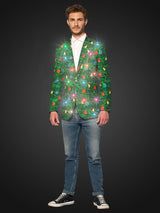 A man stands wearing the Suitmeister Christmas Green Tree Jacket Light Up, Blazer with light, which features vibrant holiday designs, paired with a white shirt, jeans, and black sneakers. He has short brown hair and a slight smile, with his hands by his sides against a dark background and the light on.
