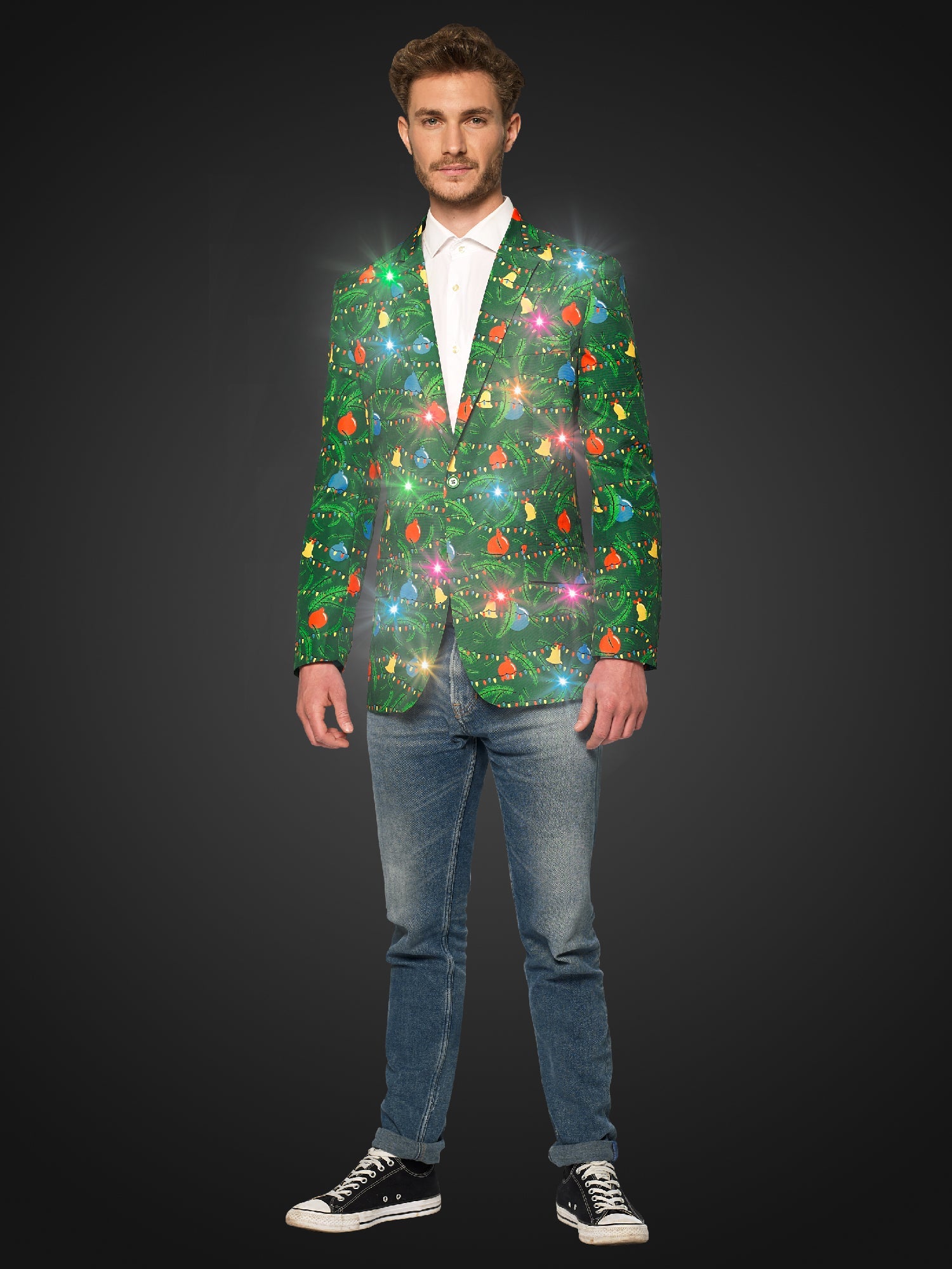 A man stands wearing the Suitmeister Christmas Green Tree Jacket Light Up, Blazer with light, which features vibrant holiday designs, paired with a white shirt, jeans, and black sneakers. He has short brown hair and a slight smile, with his hands by his sides against a dark background and the light on.