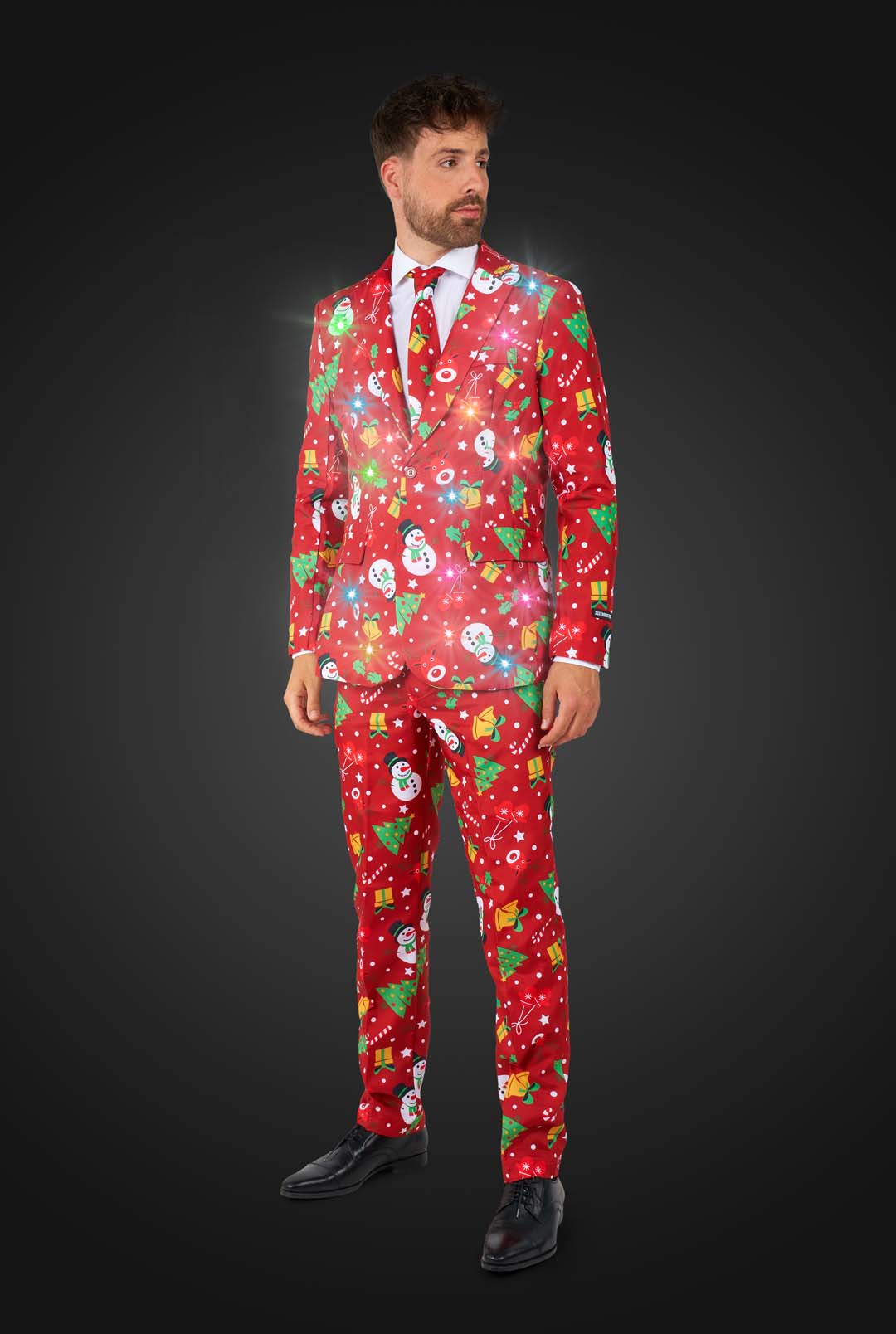 A smiling man dressed in the Suitmeister "Christmas Red Icons - Light Up" suit, featuring festive patterns like snowmen, trees, and ornaments, poses against a dark background. He adjusts his sleeve while looking at the camera. With the lights on.