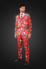 A smiling man dressed in the Suitmeister "Christmas Red Icons - Light Up" suit, featuring festive patterns like snowmen, trees, and ornaments, poses against a dark background. He adjusts his sleeve while looking at the camera. With the lights on.