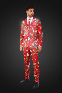 A smiling man dressed in the Suitmeister "Christmas Red Icons - Light Up" suit, featuring festive patterns like snowmen, trees, and ornaments, poses against a dark background. He adjusts his sleeve while looking at the camera. With the lights on.