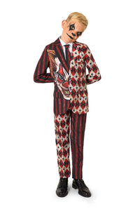 Full-body view of a child in a red and black patterned clown suit with a white shirt and black tie, standing with hands behind their back.