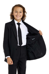 A young person with long hair, smiling, is wearing Suitmeisters Solid Black suit paired with a white shirt and black tie, standing against a white background.