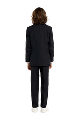 A young person with long hair, smiling, is wearing Suitmeisters Solid Black suit paired with a white shirt and black tie, standing against a white background.
