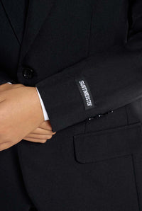 A young person with long hair, smiling, is wearing Suitmeisters Solid Black suit paired with a white shirt and black tie, standing against a white background.