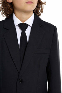 A young person with long hair, smiling, is wearing Suitmeisters Solid Black suit paired with a white shirt and black tie, standing against a white background.