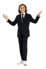 A young person with long hair, smiling, is wearing Suitmeisters Solid Black suit paired with a white shirt and black tie, standing against a white background.
