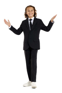 A young person with long hair, smiling, is wearing Suitmeisters Solid Black suit paired with a white shirt and black tie, standing against a white background.