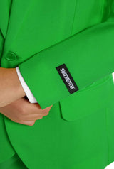 A young person with wavy hair smiles at the camera, wearing a Suitmeister Solid Green suit, white shirt, and green tie. They stand against a white background with one hand in their pocket.