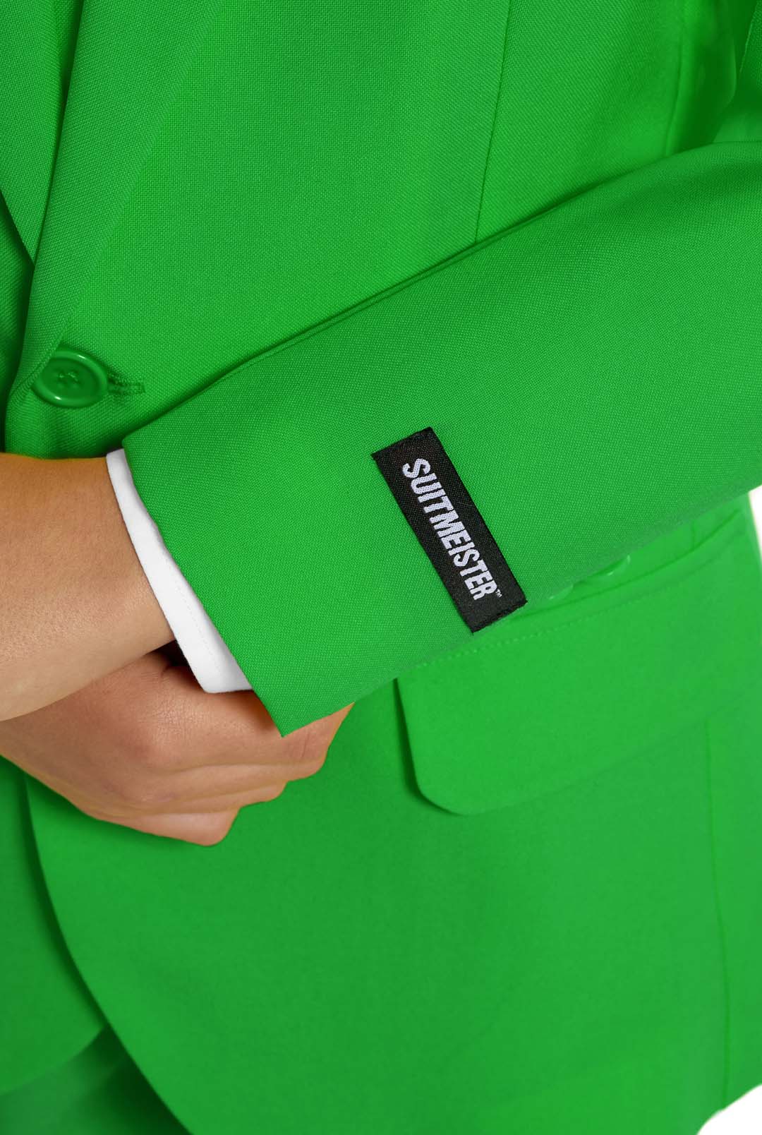 A young person with wavy hair smiles at the camera, wearing a Suitmeister Solid Green suit, white shirt, and green tie. They stand against a white background with one hand in their pocket.