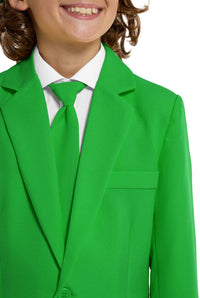 A young person with wavy hair smiles at the camera, wearing a Suitmeister Solid Green suit, white shirt, and green tie. They stand against a white background with one hand in their pocket.