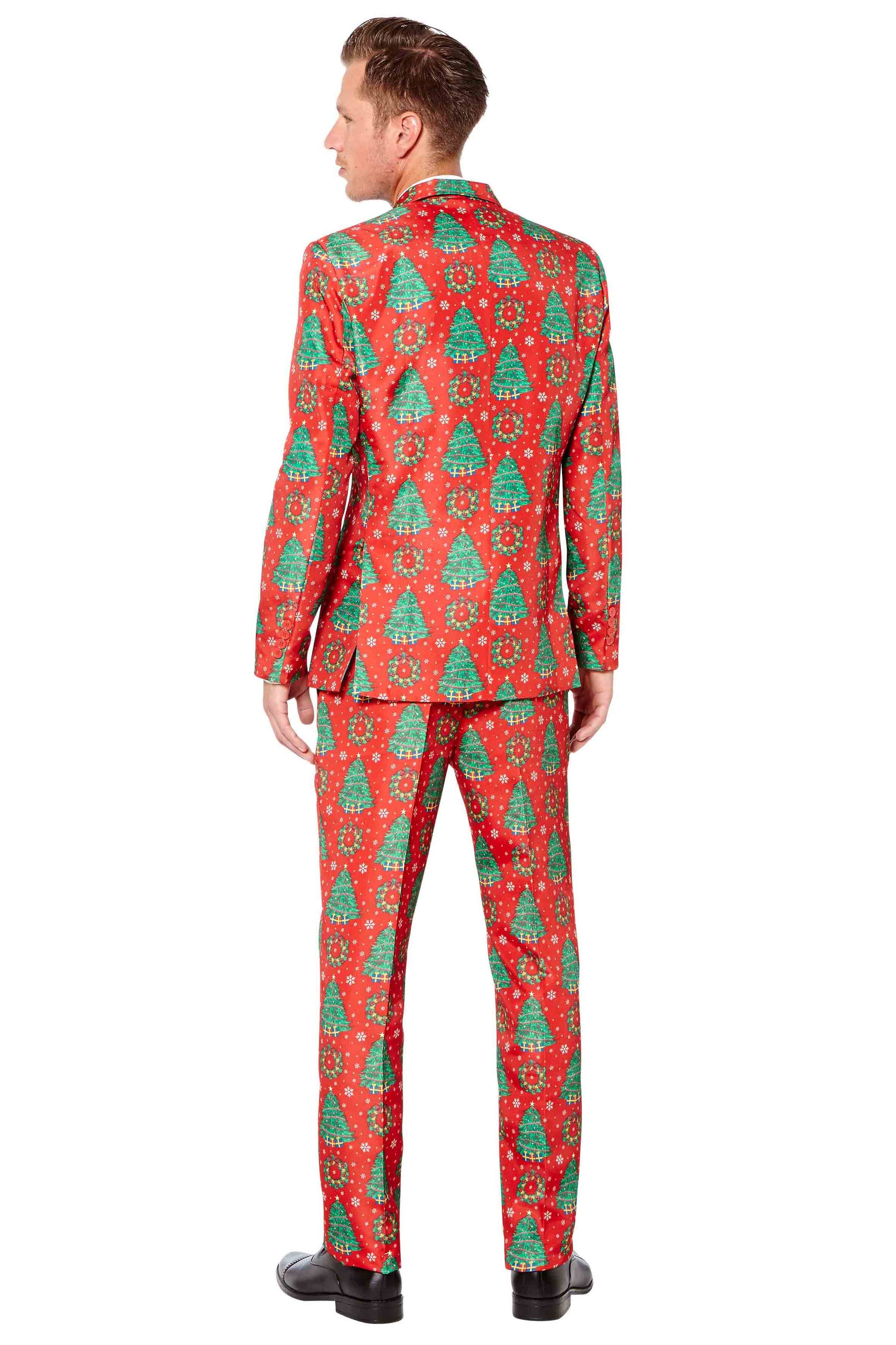 A person is wearing the Suitmeister "Christmas Trees" suit, featuring festive red fabric adorned with green Christmas tree patterns. They are standing with their back turned and have short hair. The outfit comprises a matching jacket and pants, paired with black shoes.