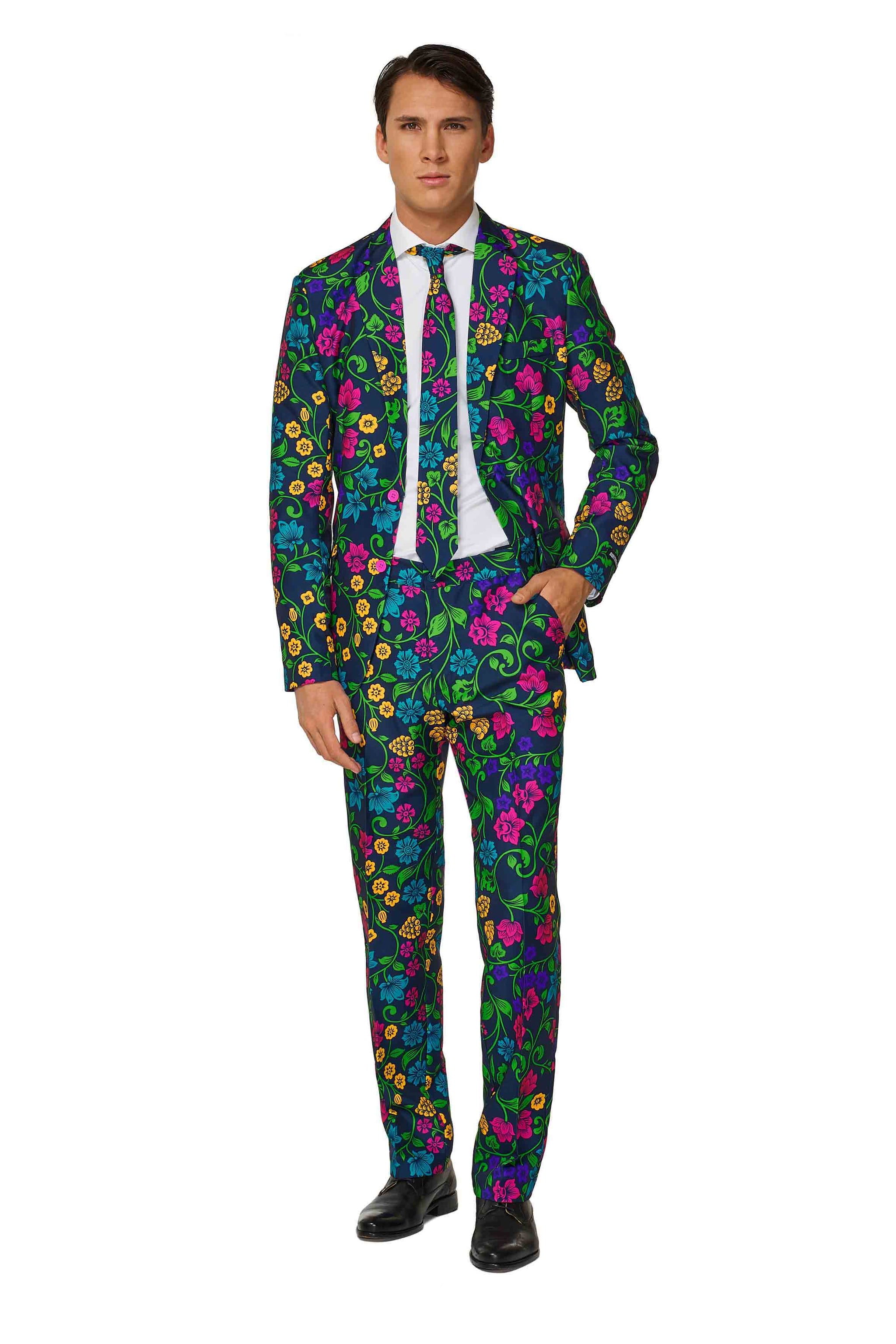 A man wearing the Suitmeister Floral, a vibrant suit adorned with a colorful floral pattern featuring blue, pink, green, and yellow flowers. He complements it with a white shirt, black tie, and black shoes while standing against a plain white backdrop.