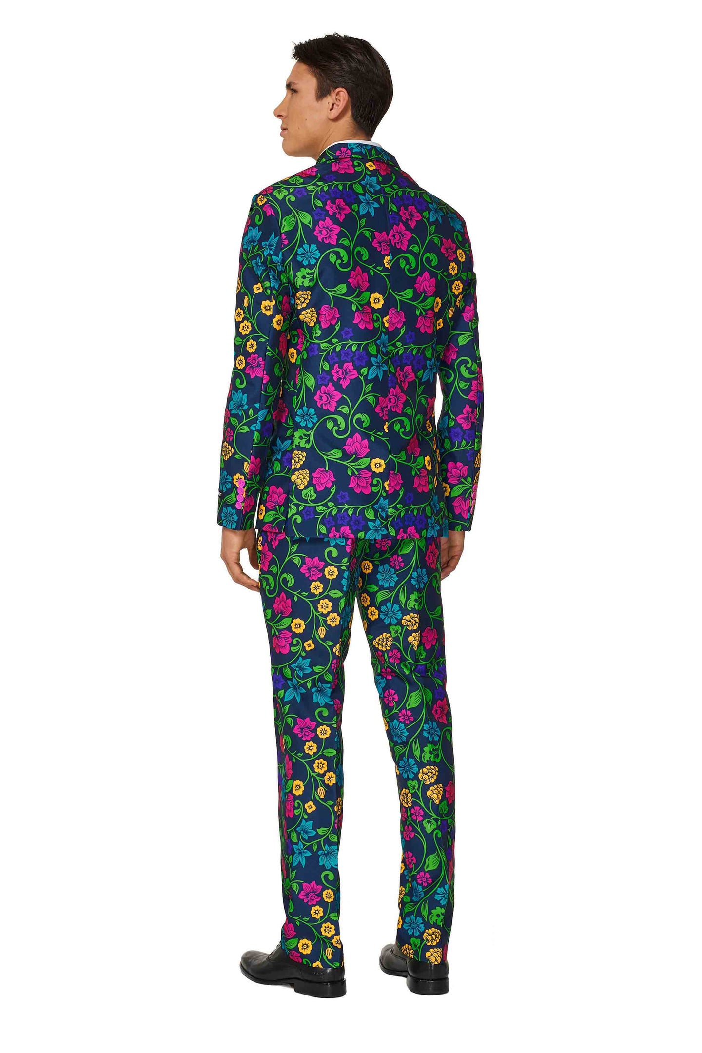 A person dressed in a vibrant Suitmeister Floral suit, characterized by a navy background adorned with bright pink, yellow, and green flowers. The individual is standing with their back to the camera and wearing black shoes.