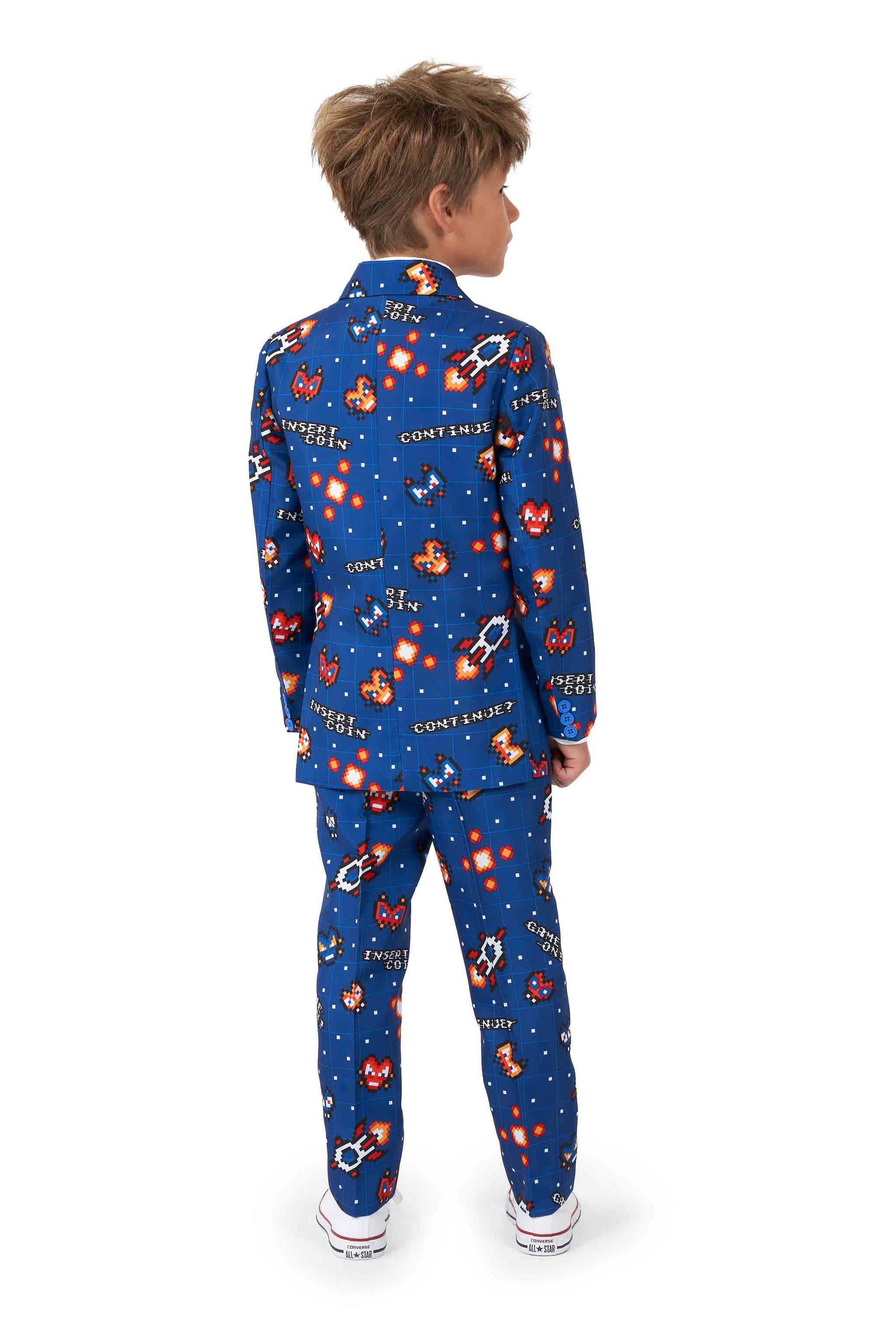 A young boy stands facing away, wearing the Retro Gamer Navy suit from Suitmeister. The outfit showcases a playful space theme with colorful patterns, including small images of rocket ships and planets, all set against cartoon graphics and texts.