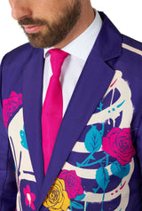 A man wearing a Suitmeister Sugar Skull Purple jacket adorned with vibrant floral and abstract designs, including roses and skeleton motifs. He pairs it with a white shirt and a vivid pink tie.
