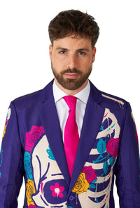 A man with a beard is wearing the Suitmeister "Sugar Skull Purple" suit jacket, decorated with colorful floral and skeleton designs. He has paired it with a white shirt and a bright pink tie, standing against a white background.