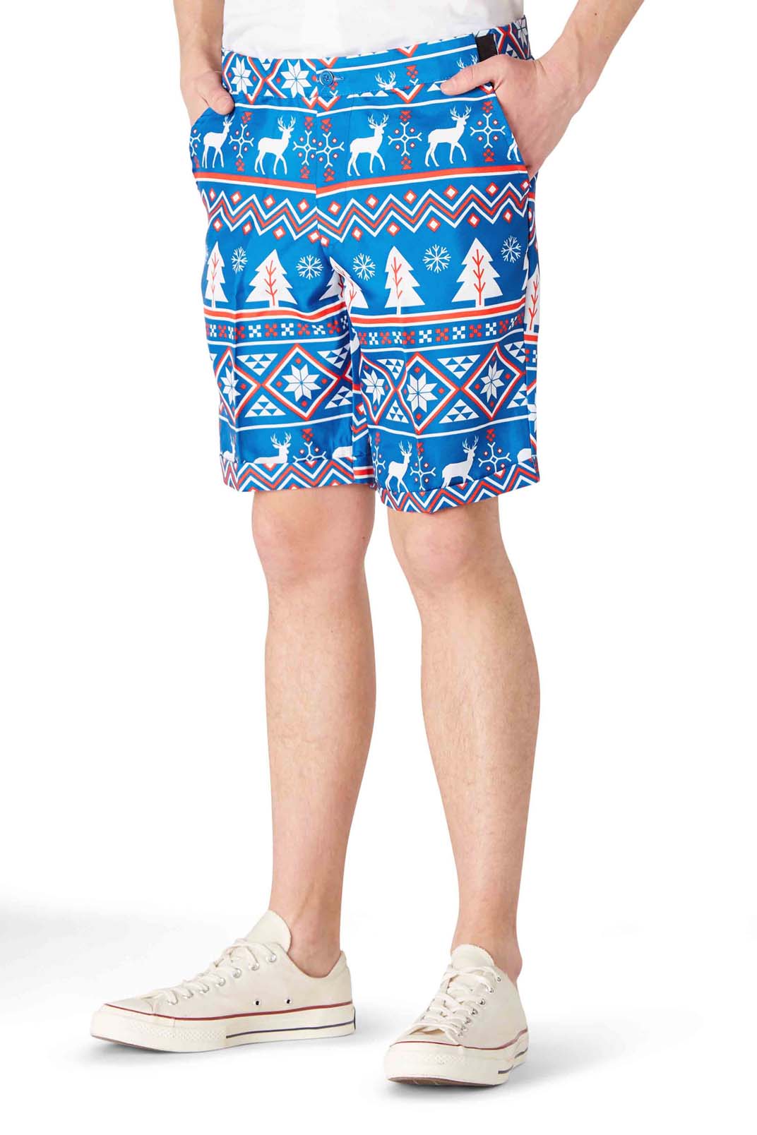 A person is wearing the "Christmas Blue Nordic" shorts by Suitmeister, featuring a festive holiday pattern with reindeer, snowflakes, and trees. They are paired with white sneakers while standing with hands in their pockets. The image highlights the shorts and lower legs.